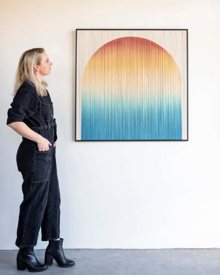 Rianne Aarts Standing In Front of Framed Orange, Yellow, and Blue Gradient Sun Piece Made From Fiber