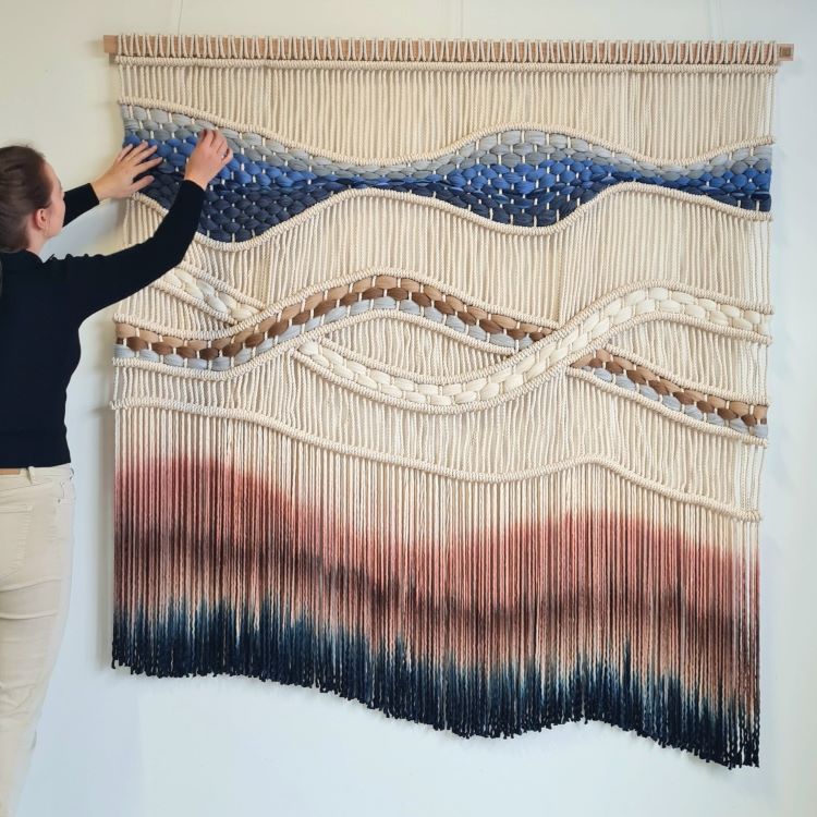 Rianne Aarts Standing In Front Of Woven Wall Hanging With Cream, Blue, And Amber Colored Thread