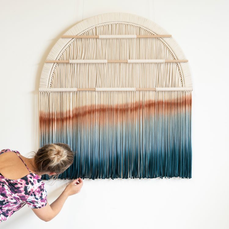 Rianne Aarts Standing In Front Of Woven Wall Hanging In Half-Moon Pattern With Amber And Blue Gradient Design At Bottom