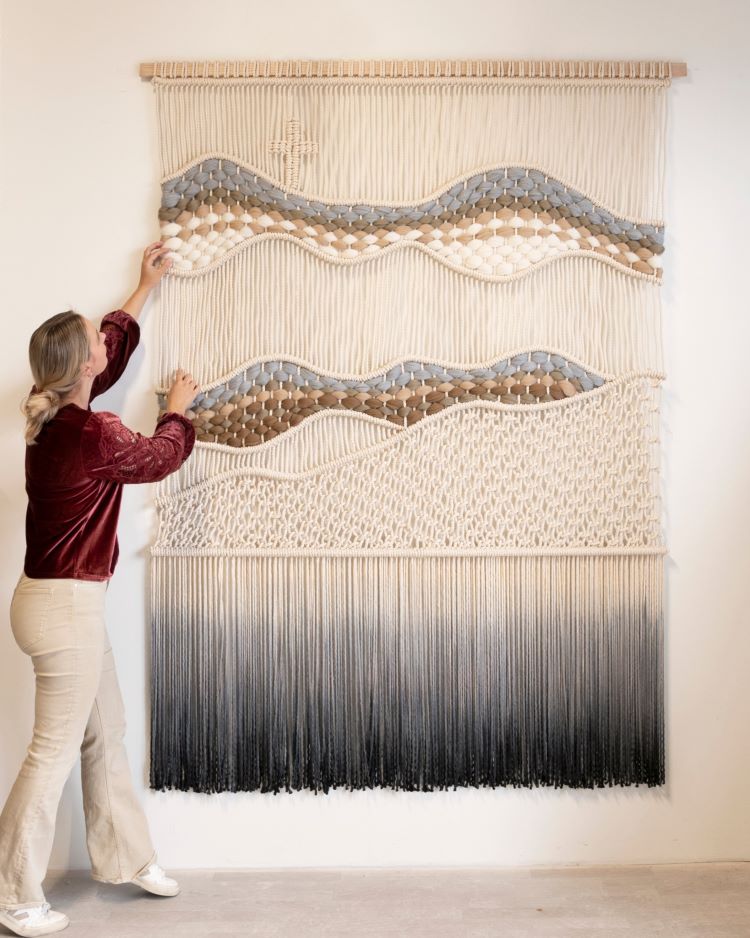 Rianne Aarts Standing In Front Of Woven Wall Hanging With Cream, Grey, And Brown Colored Thread